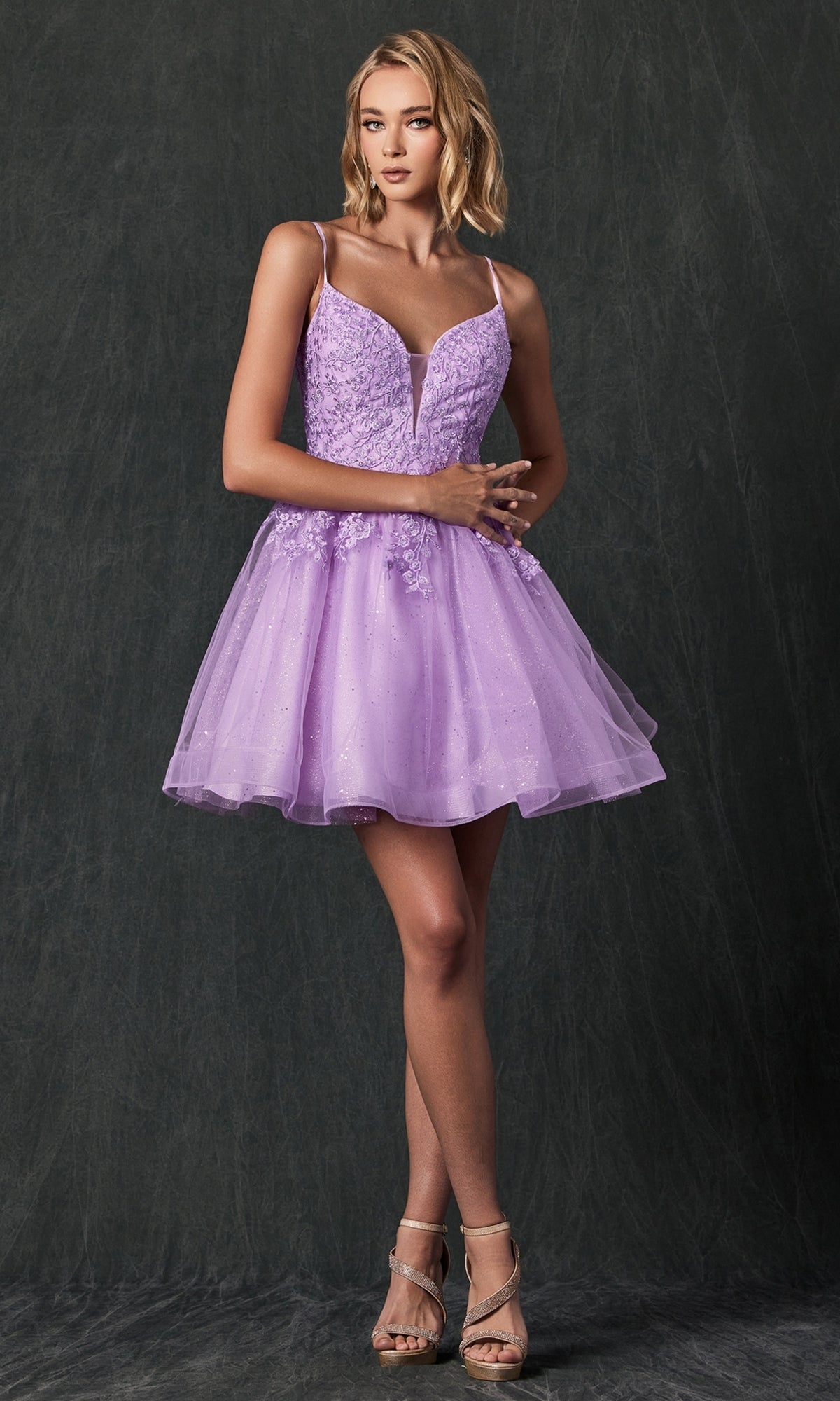 A woman models the Short Lace-Up Babydoll Homecoming Dress 859, featuring a sleeveless lavender design with a flared tulle skirt and floral details, standing against a dark textured background.